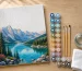 Partly finished paint by numbers canvas of a mountain lake beside 24 numbered acrylic paint pots and three brushes on a wooden table